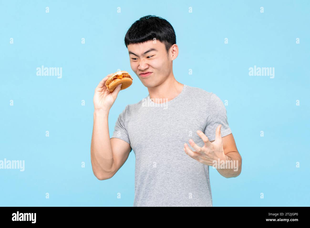 Nervöser asiatischer Mann in Freizeitkleidung, der frischen ungesunden Burger in der Hand betrachtet, während er auf hellblauem isoliertem Hintergrund im Studio steht Stockfoto