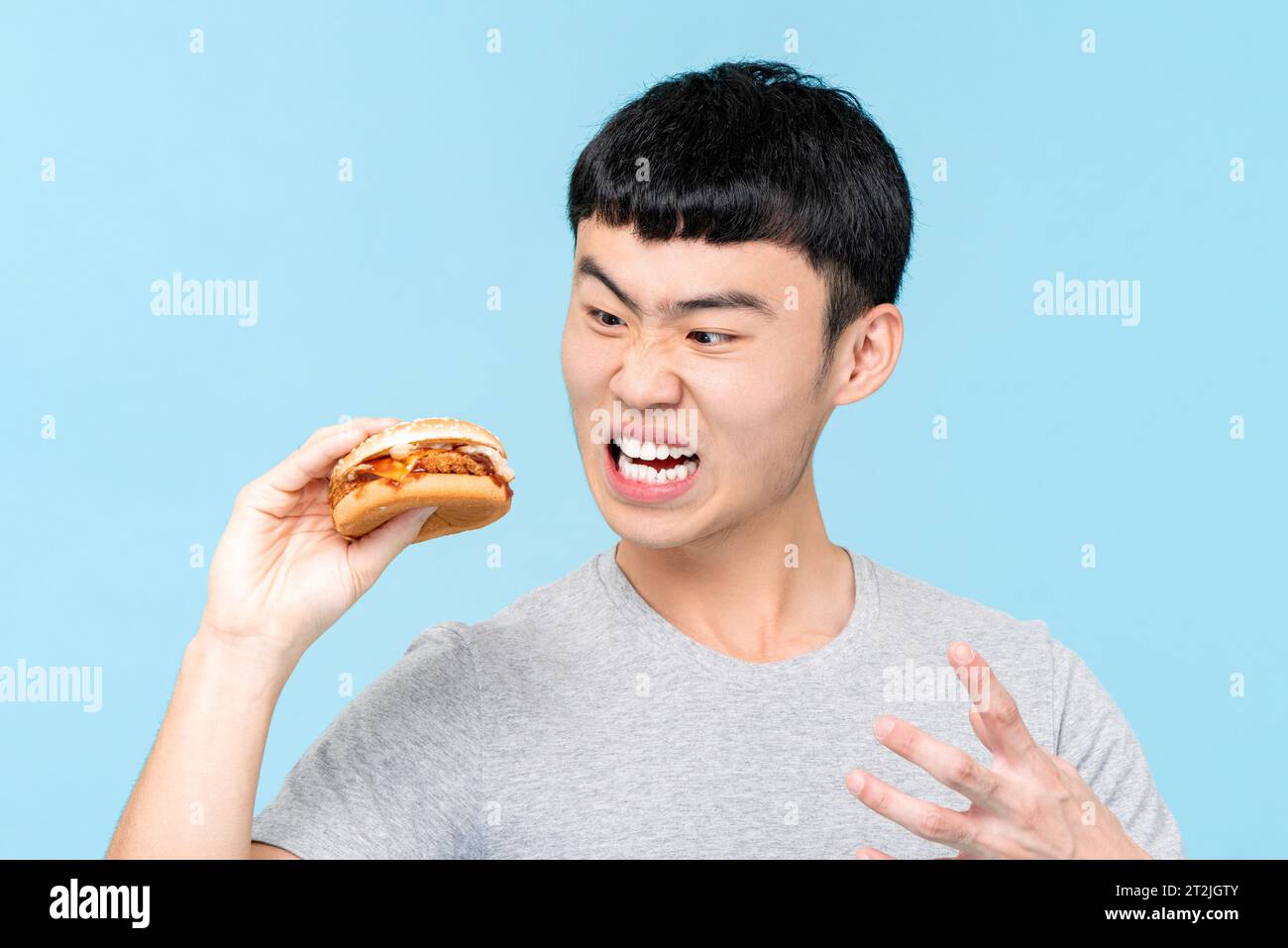 Wütender asiatischer Mann auf Diät, der ungesunde Burger betrachtet, während er auf hellblauem isoliertem Hintergrund im Studio steht Stockfoto