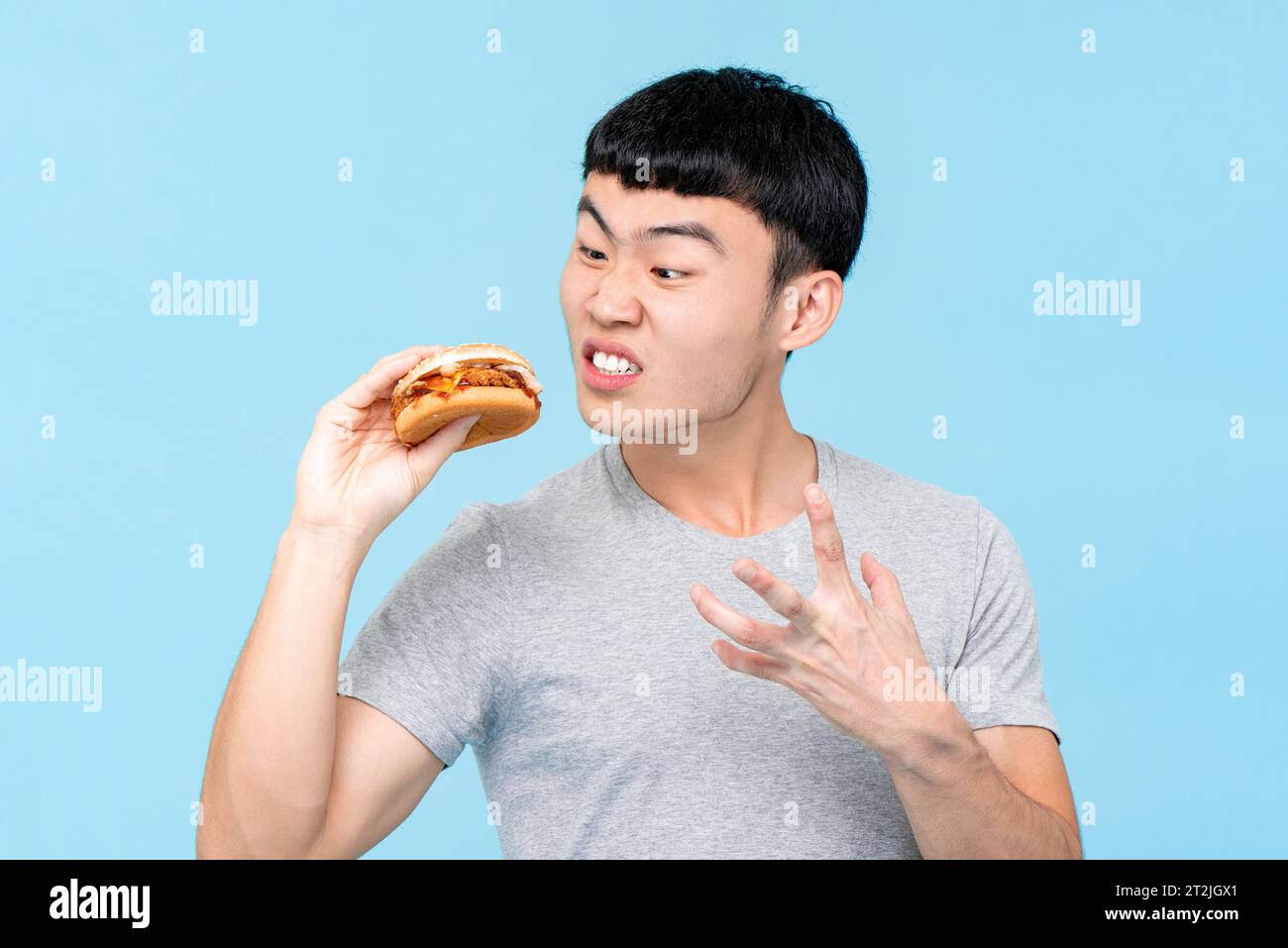 Wütender asiatischer Mann, der auf frischen ungesunden Burger blickt, während er auf hellblauem isoliertem Hintergrund im Studio steht Stockfoto