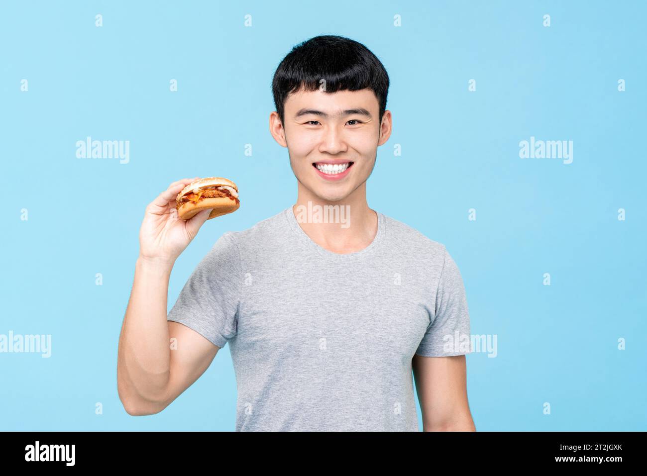 Positiver asiatischer Mann im lässigen Outfit, der leckeren frischen Burger mit einem Lächeln auf hellblauem isoliertem Hintergrund hält Stockfoto