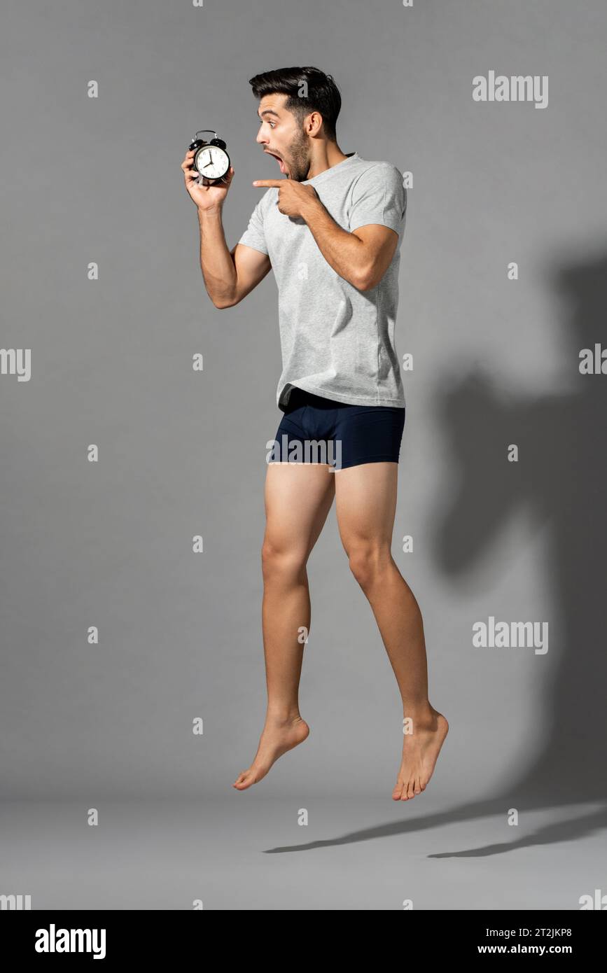 Schockierter kaukasischer Mann mit Wecker in der Hand, der springt, nachdem er spät auf grauem, isoliertem Hintergrund im Studiozimmer aufgewacht ist Stockfoto