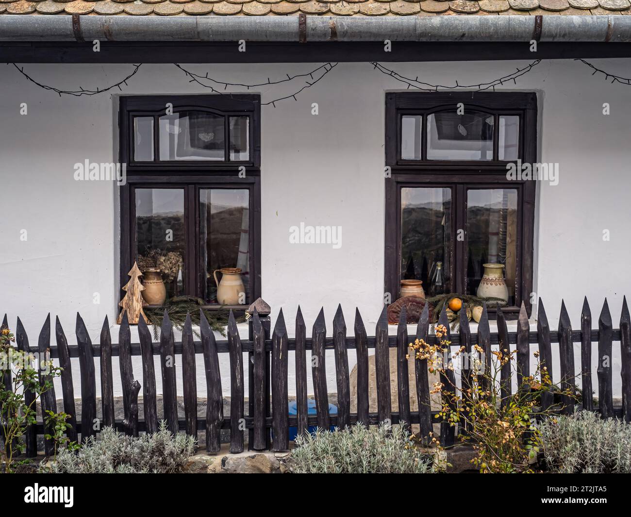 Frontale Detailansicht eines traditionellen Dorfhauses mit weißen Wänden, Holzfenstern und Zaun, Töpfen und Pflanzen als Dekoration, Holloko, Ungarn, Ostern Stockfoto