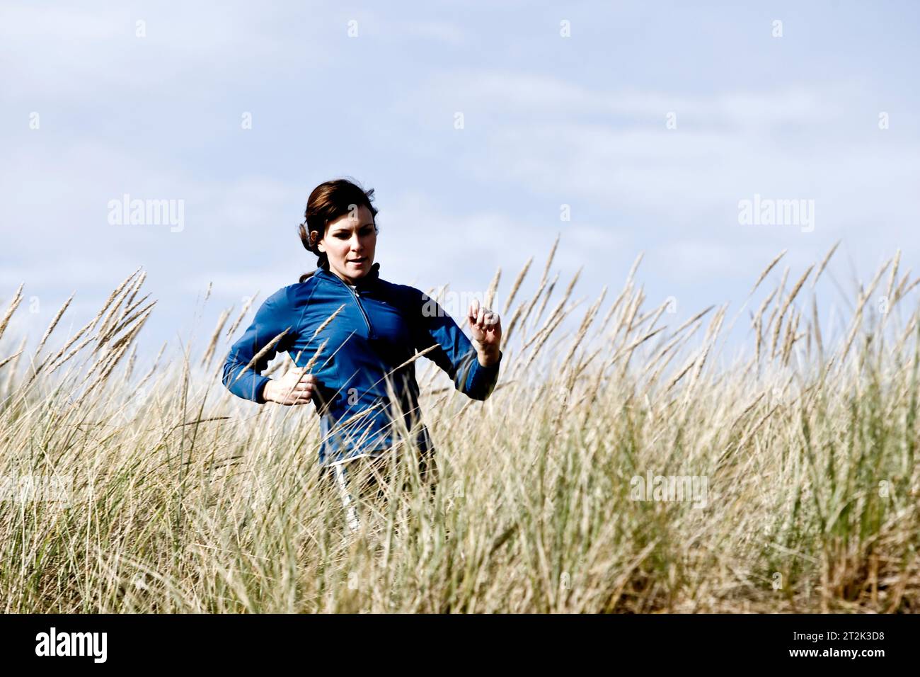 Eine Frau, die auf einem Sandweg mit hohem Gras am Meer läuft. Stockfoto
