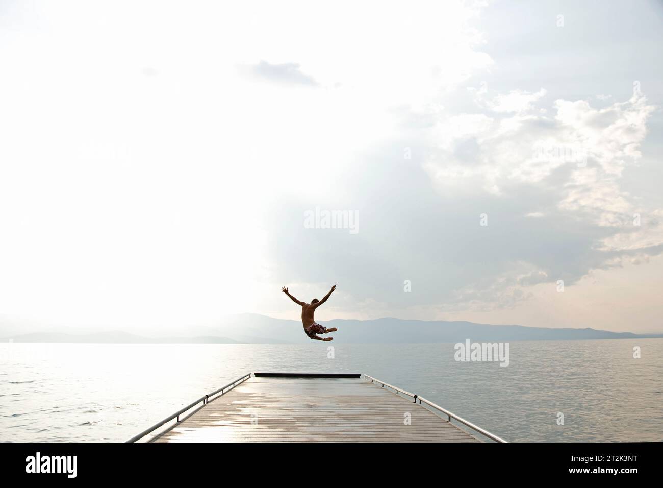 Ein junger Teenager wirft seine Arme und tritt mit den Beinen in die Luft, während er von einem Dock springt, um im kühlen Wasser des Flathead Lake, Montana, zu schwimmen. Stockfoto