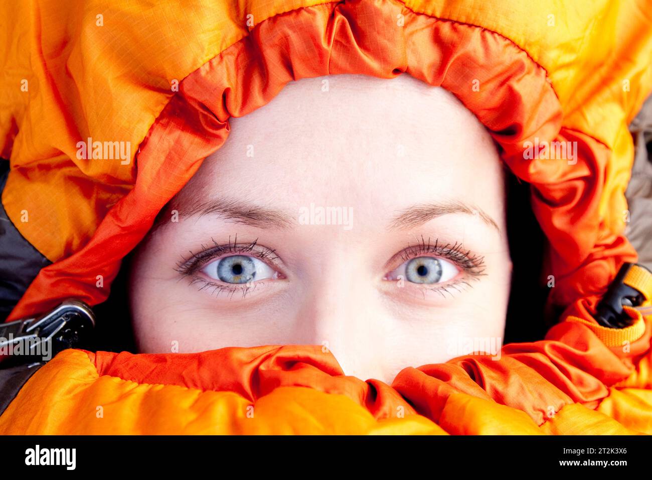 Eine Frau mit hellblauen Augen blickt direkt in die Kamera, während sie fest in ihrem orangefarbenen Schlafsack sitzt. Stockfoto