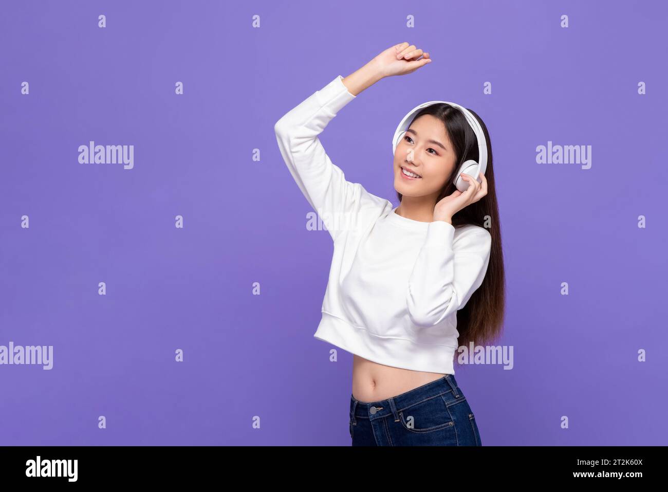 Fröhliche glückliche lächelnde asiatische Frau, die Musik auf Kopfhörern hört und im lila Farbstudio im isolierten Hintergrund tanzt Stockfoto