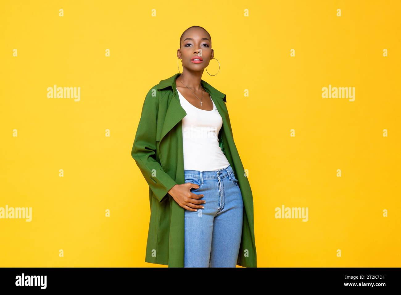 Porträt einer stilvollen, selbstbewussten afroamerikanerin der Millennials mit einem rasierten Kopf, der einen Mantel trägt und vor einem isolierten gelben Hintergrund posiert Stockfoto