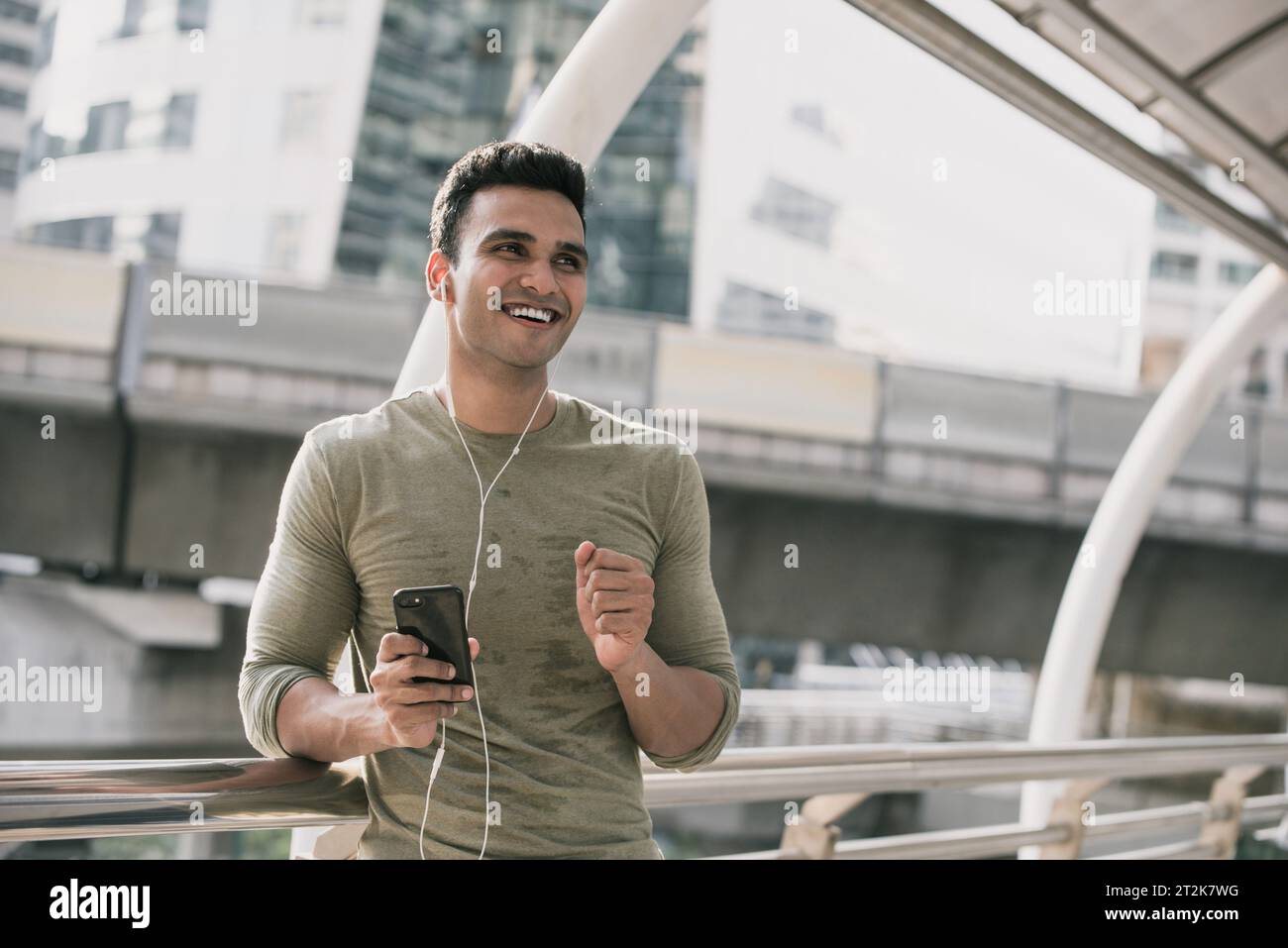 Glücklicher, gutaussehender Indianer, der Ohrhörer trägt und Musik auf dem Smartphone in der Stadt hört Stockfoto