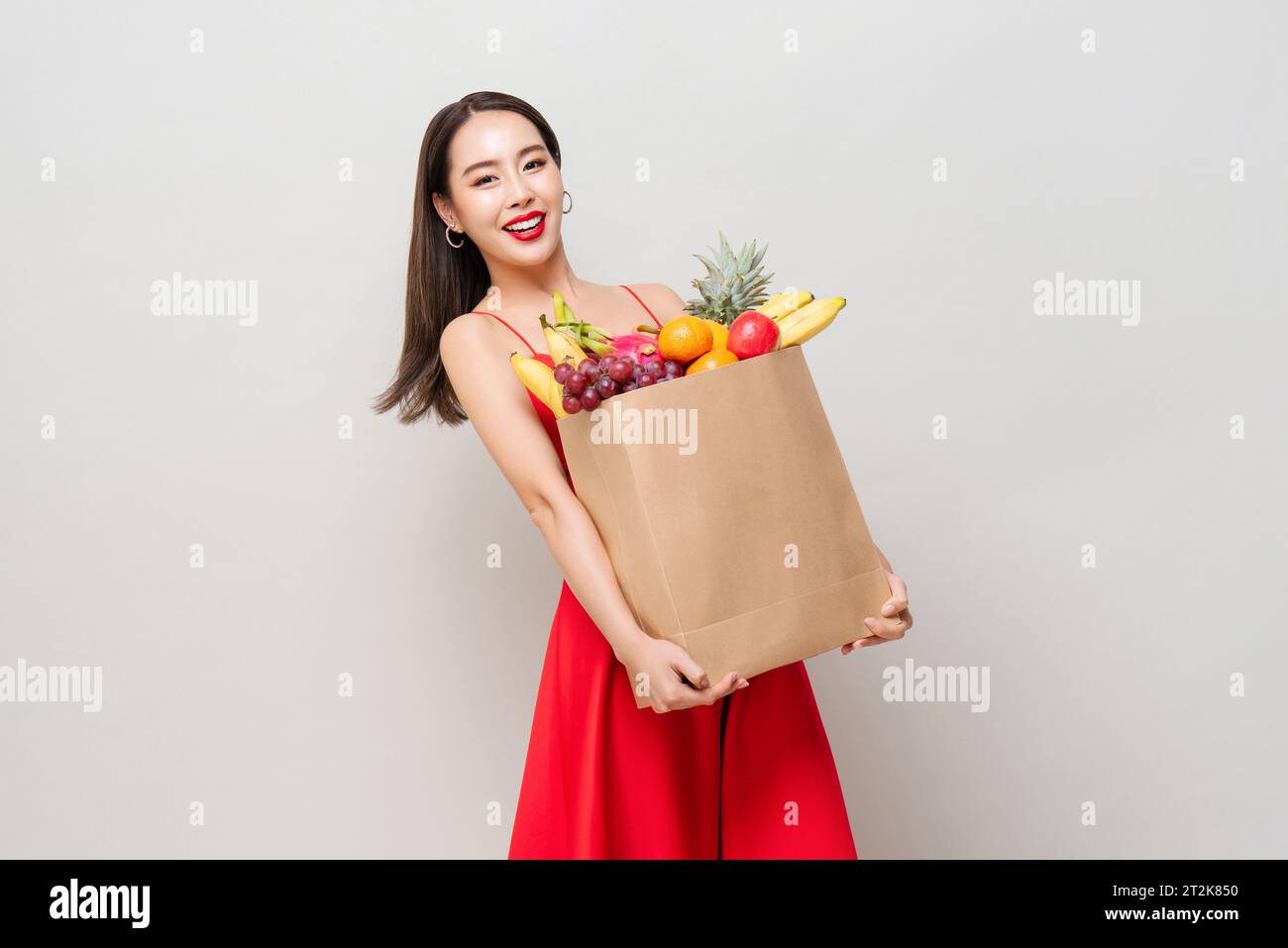 Glückliche asiatische Frau in rotem Kleid, die Papier-Lebensmittelbeutel voller tropischer Früchte in grauem Studio isoliert Hintergrund hält Stockfoto