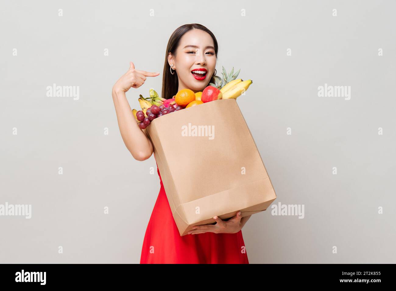 Lächelnde asiatische Frau in rotem Kleid, die Papier-Lebensmittelbeutel voller tropischer Früchte in grauem Studio isoliert Hintergrund hält Stockfoto