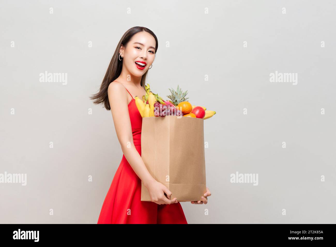 Lächelnde asiatische Frau in rotem Kleid, die Papier-Lebensmittelbeutel voller tropischer Früchte in grauem Studio isoliert Hintergrund hält Stockfoto