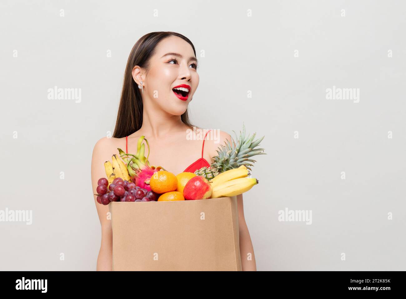 Überraschte asiatische Frau, die Papiertüte voller tropischer Früchte hält, die aufblickte, um Raum in grauem Studio isoliertem Hintergrund zu kopieren Stockfoto