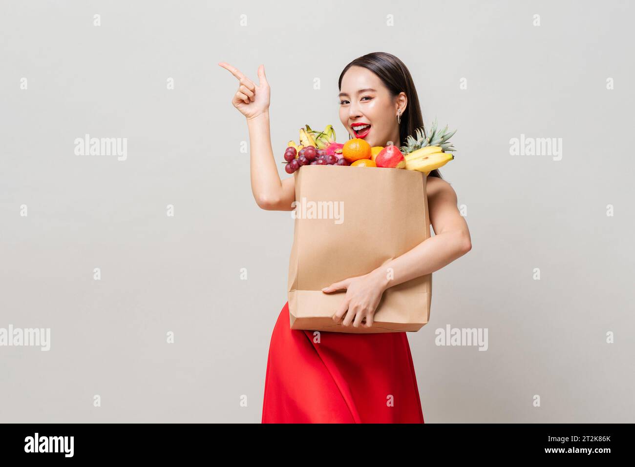 Hübsche asiatische Frau in rotem Kleid, die Papiertüte voller tropischer Früchte hält, die Hand nach oben zeigt in grauem Studio isoliertem Hintergrund Stockfoto