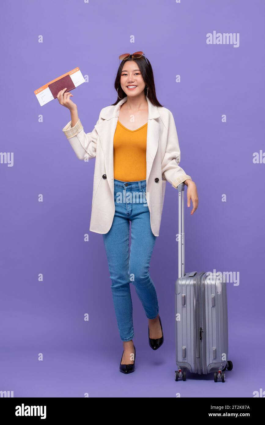 Schöne junge asiatische Touristenfrau mit Gepäck, die Bordkarte und Reisepass zeigt, die bereit sind, zu fliegen, isoliert auf lila Hintergrund Stockfoto