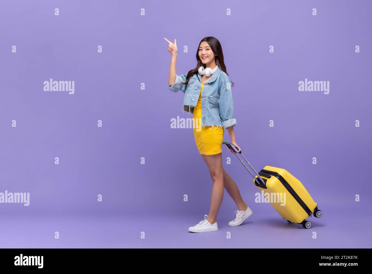 Ganzkörperporträt der jungen hübschen asiatischen Frau Touristen mit Koffer zeigt Finger nach oben in lila Farbe Studio isolierten Hintergrund Stockfoto