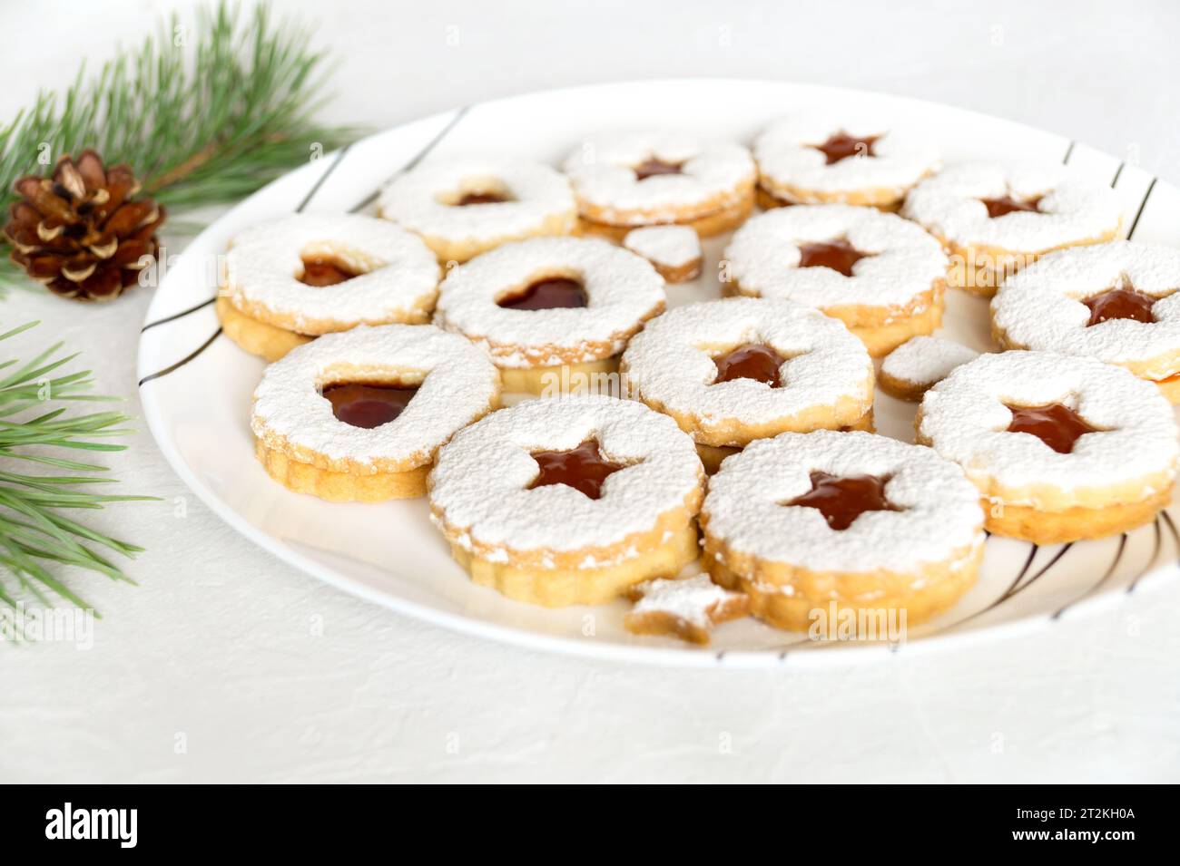 Selbstgemachte Weihnachtskekse mit Marmelade, mit Puderzucker bestreut auf weißem Teller. Weihnachtliche Atmosphäre mit Tannenzweigen und Tannenzapfen Stockfoto