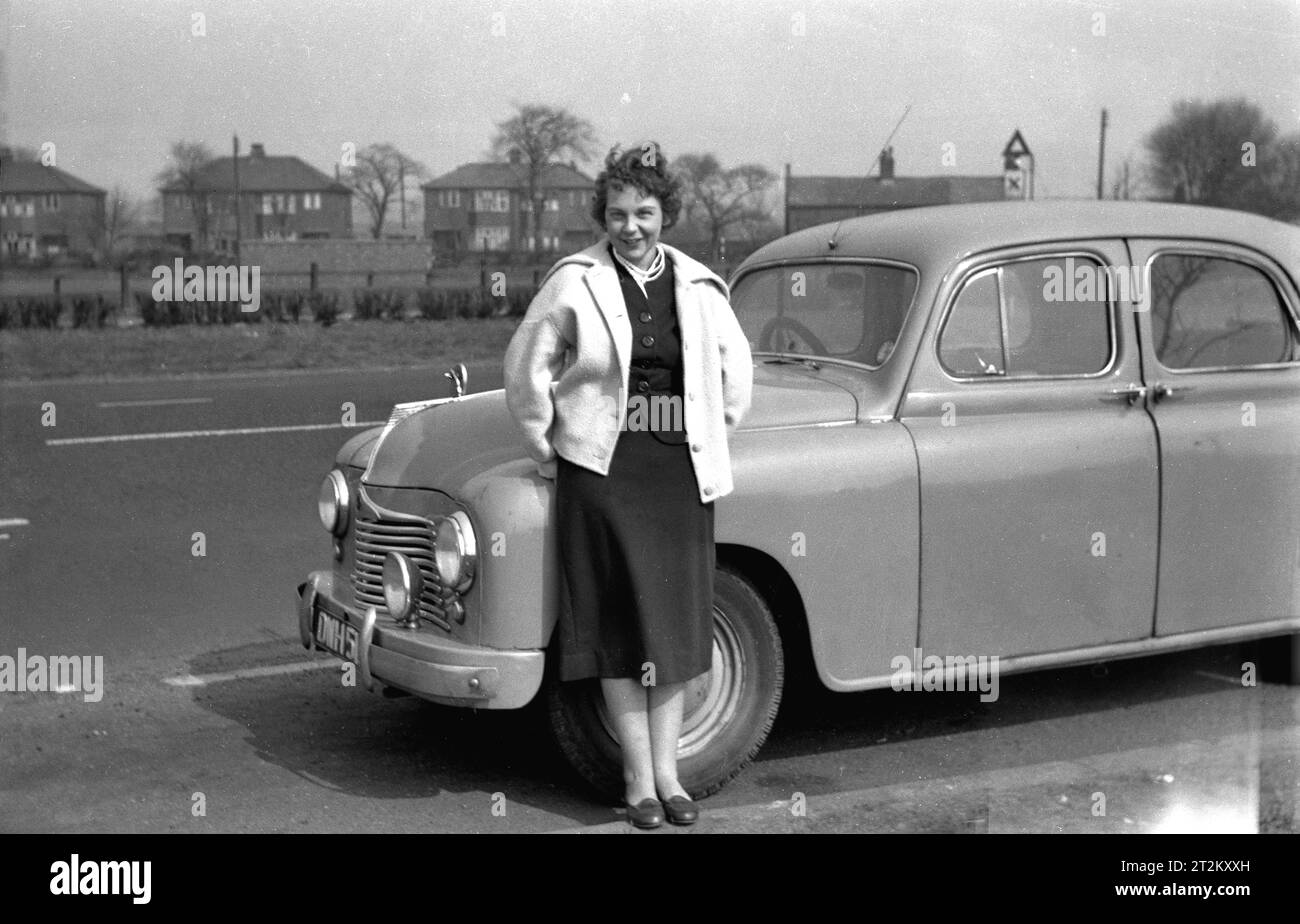 1960er Jahre, historisch, draußen, parkte neben einer Vorstadtstraße, eine Dame, die für ein Foto bei ihrem Auto aus der Zeit stand. England, Großbritannien. Beachten Sie den Griff an den Hintertüren des Fahrzeugs neben dem Griff der Vordertür, d. h. die Türen waren nach hinten aufklappbar und so nach hinten geöffnet. Hecktüren waren bei Autos, die Anfang des 20. Jahrhunderts hergestellt wurden, üblich, aber da solche Autos keine Sicherheitsgurte hatten, wurden sie als weniger sicher angesehen als eine Vordertür, da die Wahrscheinlichkeit, beim Öffnen des Fahrzeugs herauszufallen, größer war. Stockfoto