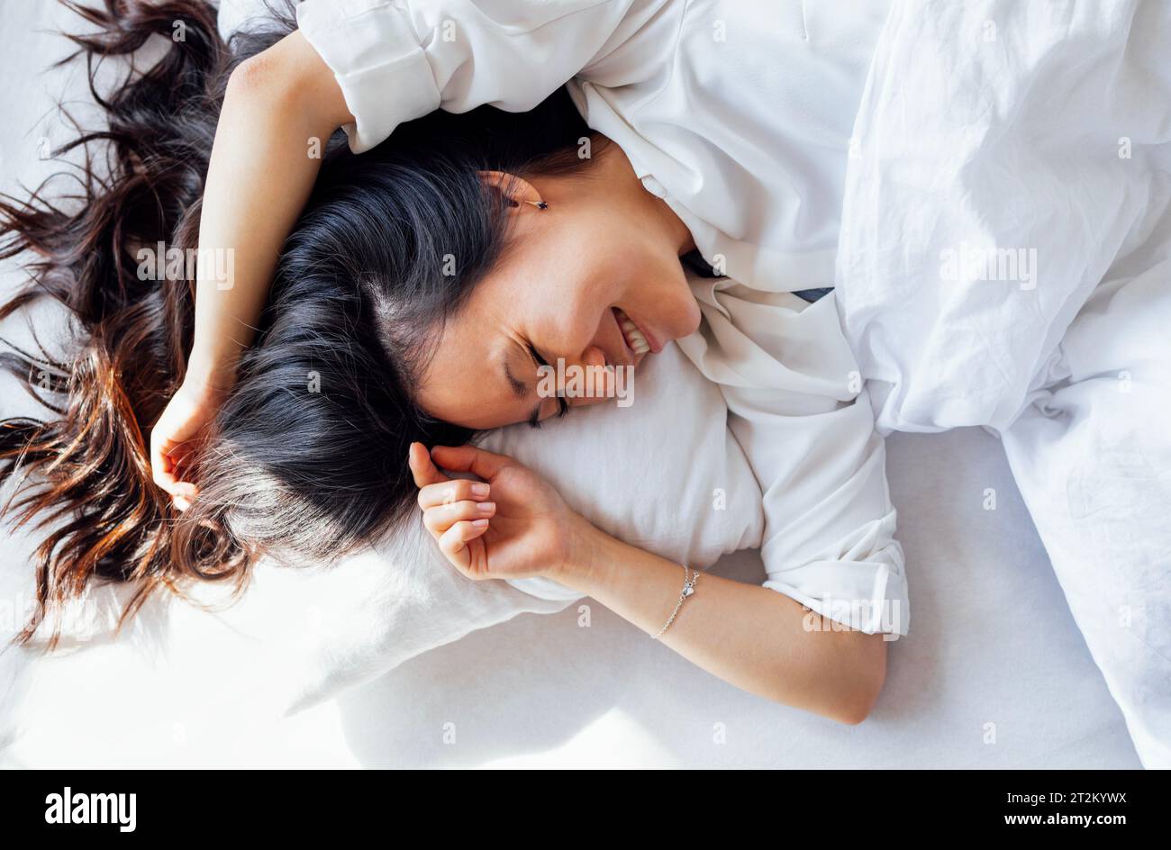 Attraktive glückliche asiatische Frau liegt morgens auf einem weißen Leinenbett. Das junge Mädchen lächelt und wacht gerne auf. Sonnenlicht auf dem Kissen und der fema Stockfoto