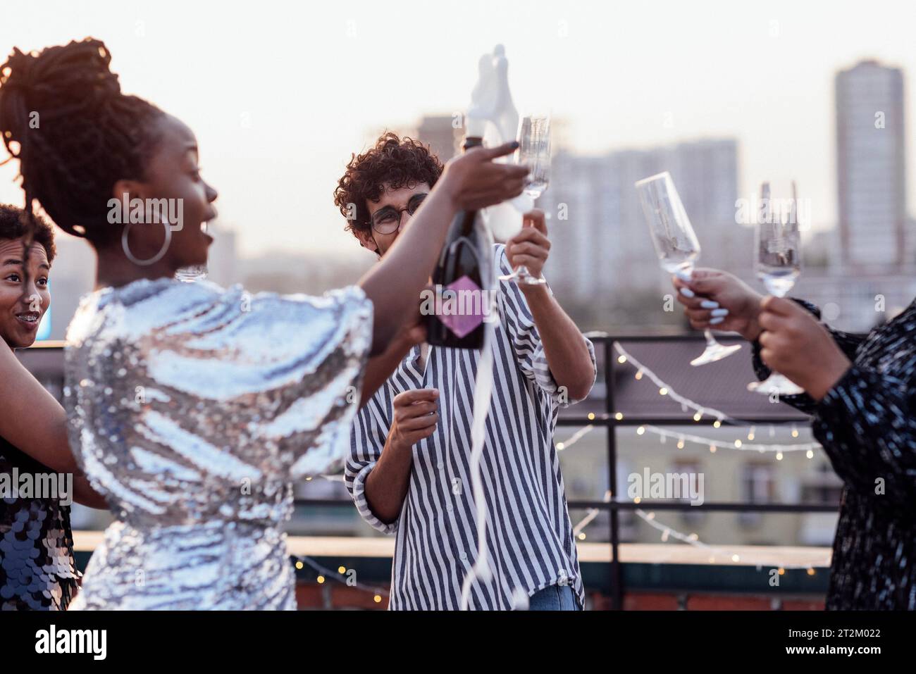 Dunkelhäutiges Mädchen gießt Champagner in Gläser ihrer multinationalen Freunde. Süße, glückliche Leute, die Spaß auf einer Dachparty haben. Afrikanische Frauen in elegantem d Stockfoto