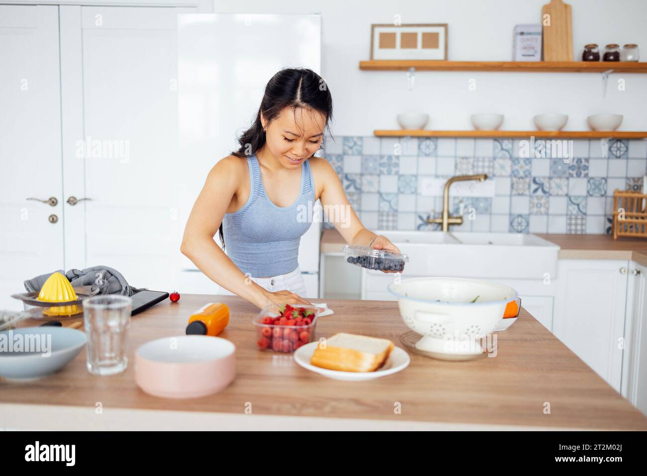Charmante asiatische Frauen bereiten Frühstück mit Beeren in der Küche zu. Eine attraktive junge Frau macht Sandwiches. Das schöne lächelnde Mädchen wischt den Tisch im Dinin ab Stockfoto