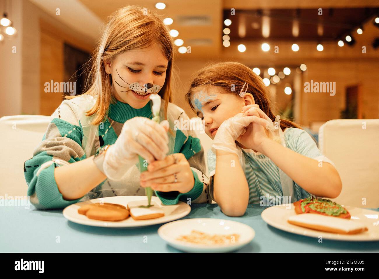 Zwei Mädchen mit Aquagrim auf dem Gesicht schmücken Weihnachtskekse mit einem Gebäckbeutel in einem Café oder Restaurant. Schwestern in legeren Kleidern haben Spaß und Vater Stockfoto