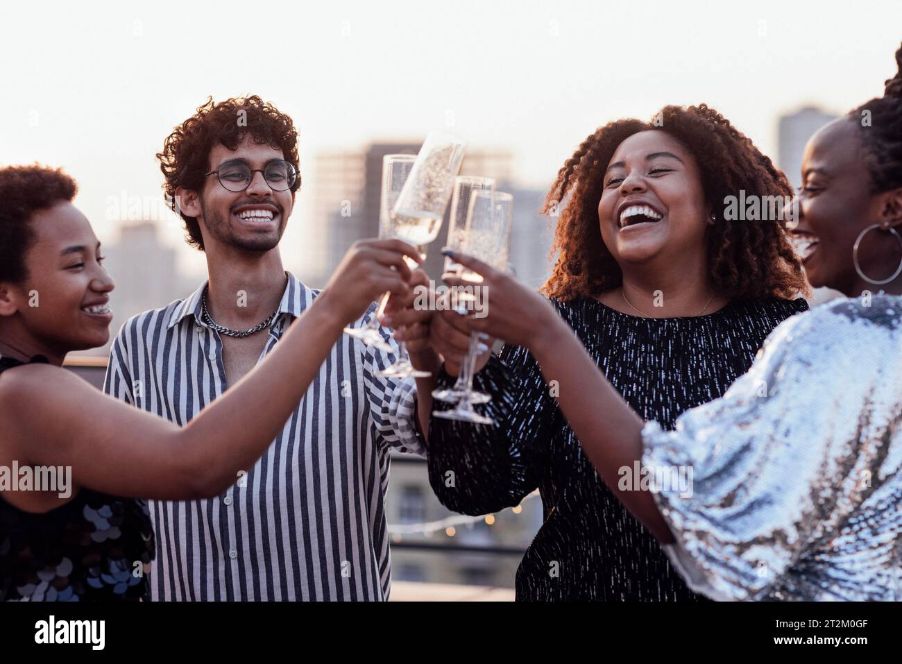 Dunkelhäutiges Mädchen gießt Champagner in Gläser ihrer multinationalen Freunde. Süße, glückliche Leute, die Spaß auf einer Dachparty haben. Afrikanische Frauen in elegantem d Stockfoto