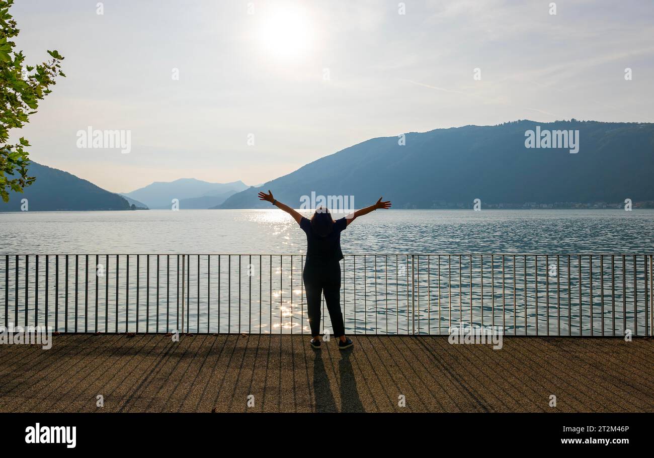 Frau mit Schatten stehend mit ausgestreckten Armen an der Uferpromenade mit Geländer zum Luganer See mit Berg an einem sonnigen Sommertag in Bissone Stockfoto