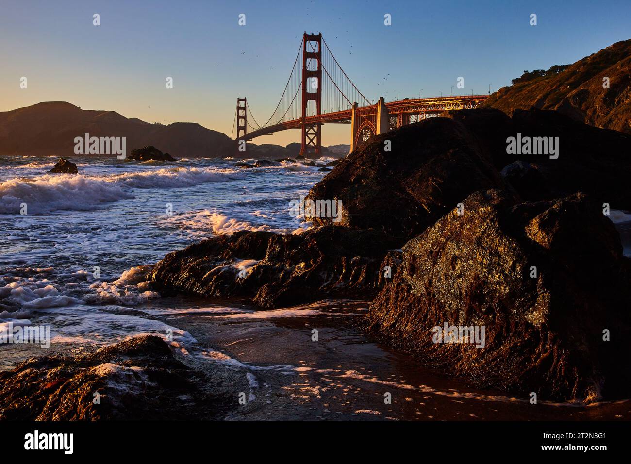 Goldenes Licht markante dunkle moosige Felsen mit brechender Welle und entfernter Golden Gate Bridge Stockfoto