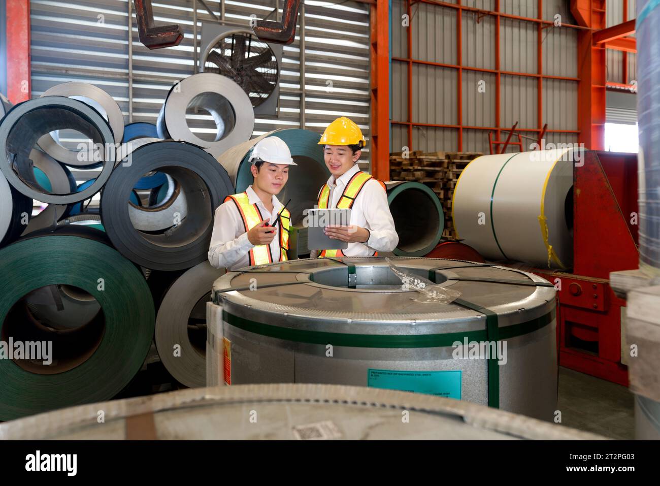 Industrielle Lagerhausszene mit zwei sicherheitsbewussten Männern, die Stahlblechrollen inspizieren, mit dem rauen Kern der täglichen manuellen Arbeit und Stockfoto