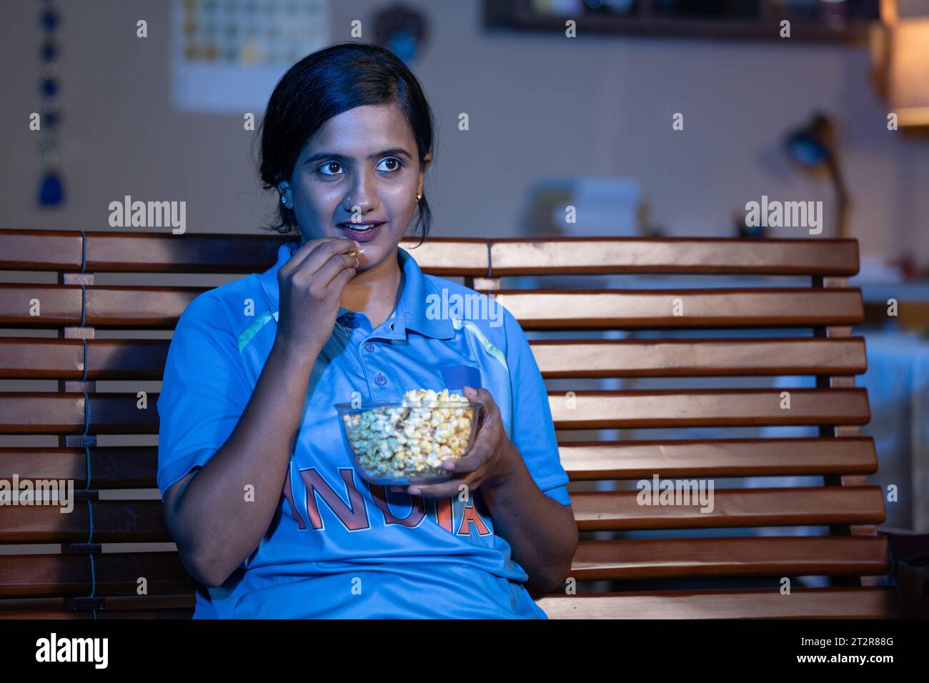 Kurioses junges Mädchen in indianertrikot, das Cricket-Match im fernsehen oder im Fernsehen zuhause beim Essen von Popcorns ansieht - Konzept von Unterstützer, Weltmeisterschaft und Stockfoto