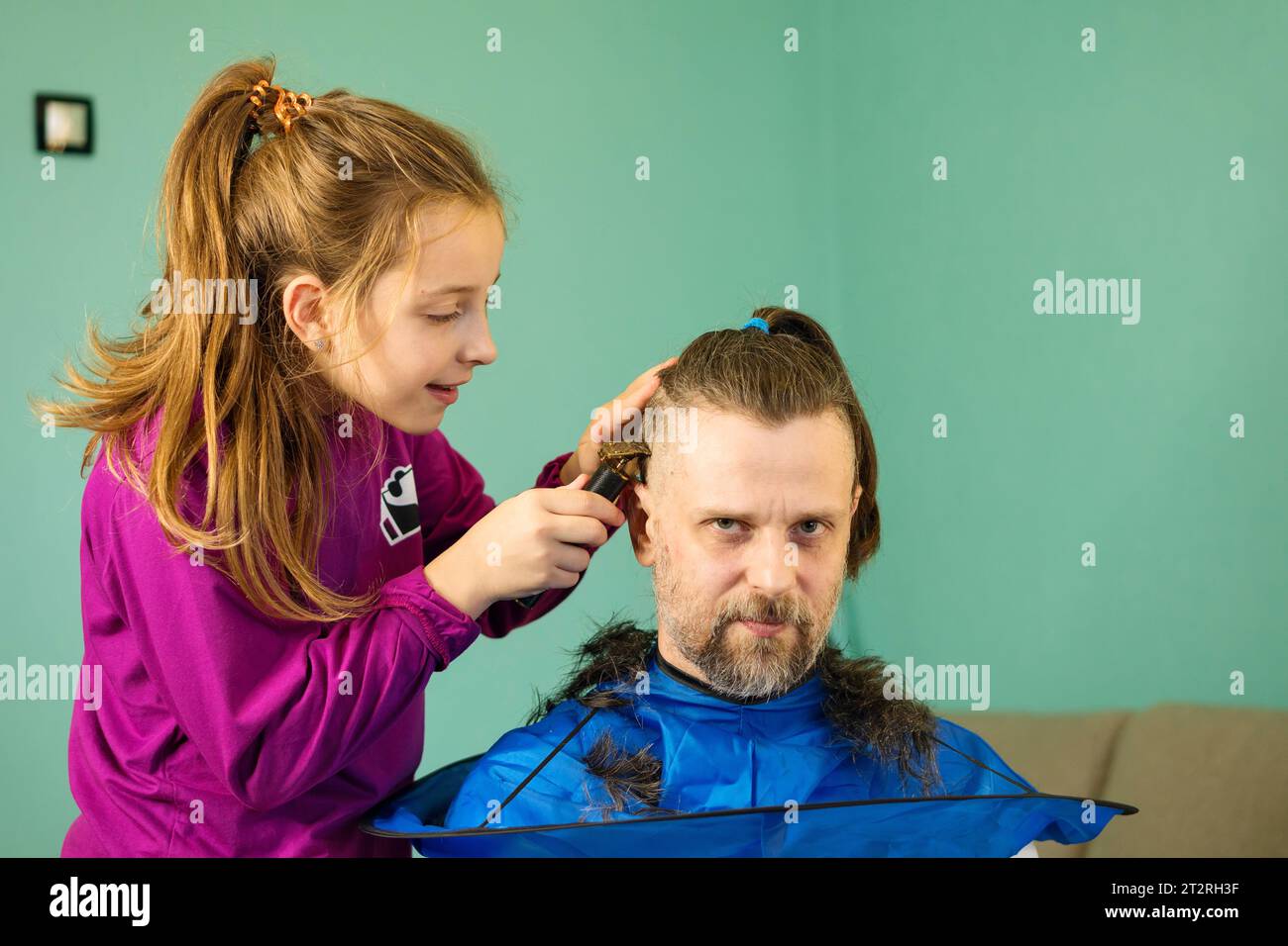 Fröhliches Mädchen schneidet zu Hause spielerisch die Haare ihres Vaters und schafft eine Bindung und Erinnerungen durch eine lustige und unschuldige Haarverwandlung. Stockfoto