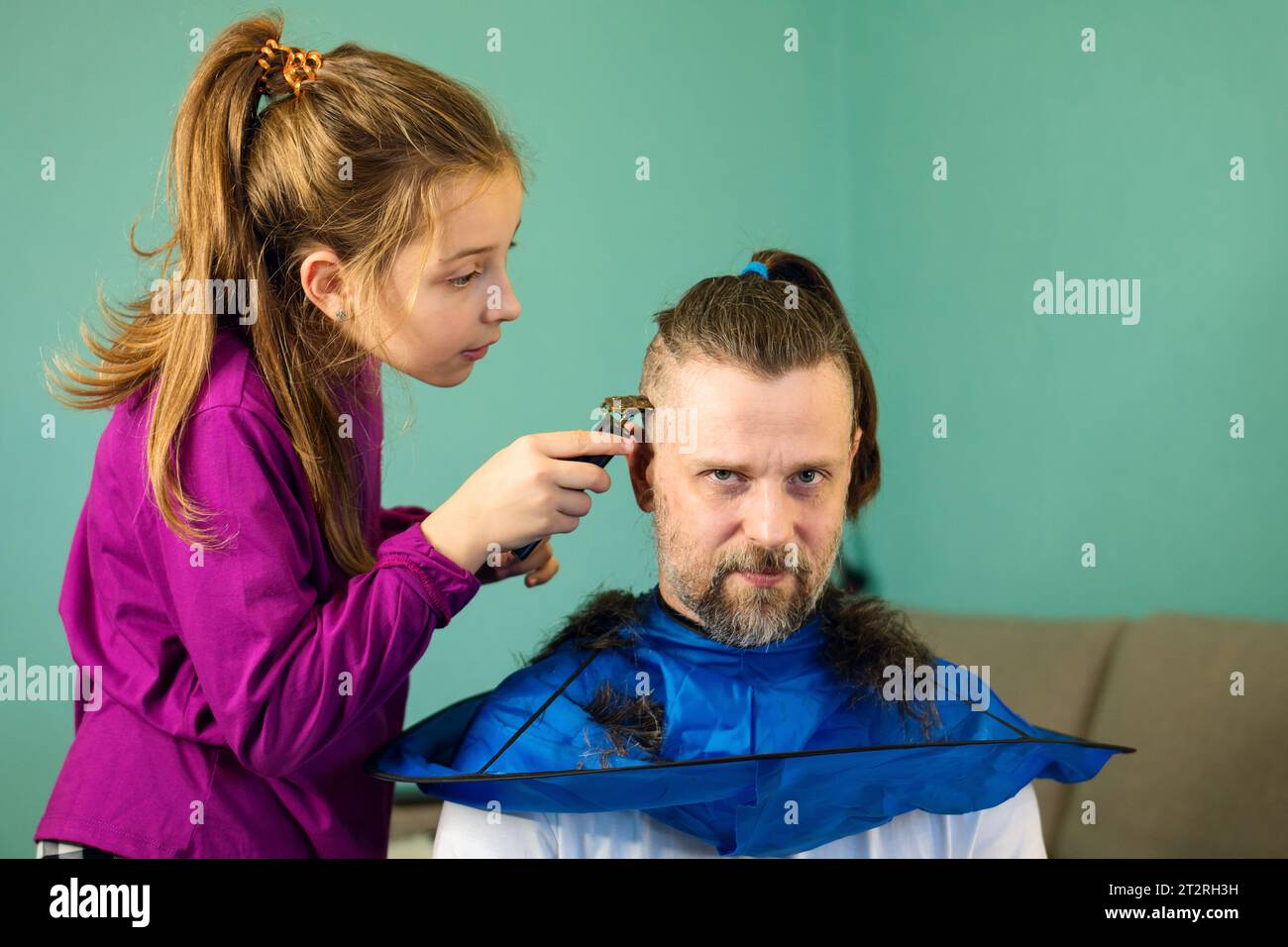 Fröhliches Mädchen schneidet zu Hause spielerisch die Haare ihres Vaters und schafft eine Bindung und Erinnerungen durch eine lustige und unschuldige Haarverwandlung. Stockfoto
