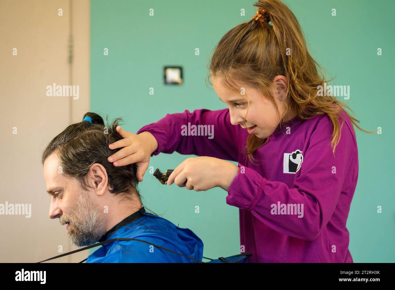 Fröhliches Mädchen schneidet zu Hause spielerisch die Haare ihres Vaters und schafft eine Bindung und Erinnerungen durch eine lustige und unschuldige Haarverwandlung. Stockfoto
