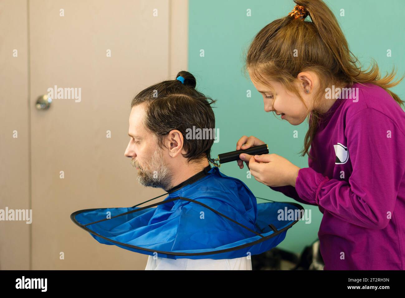 Fröhliches Mädchen schneidet zu Hause spielerisch die Haare ihres Vaters und schafft eine Bindung und Erinnerungen durch eine lustige und unschuldige Haarverwandlung. Stockfoto
