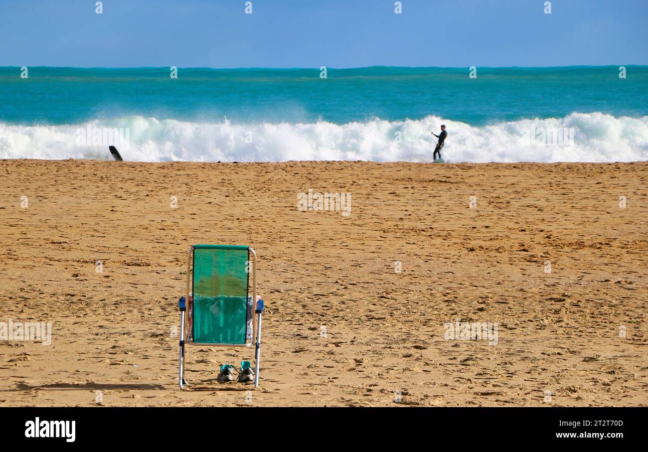 Grüner Sessel mit Schuhen darunter an einem einsamen Strand und weit entfernten Surfern mit rauem Meer Sardinero Santander Kantabrien Spanien Stockfoto