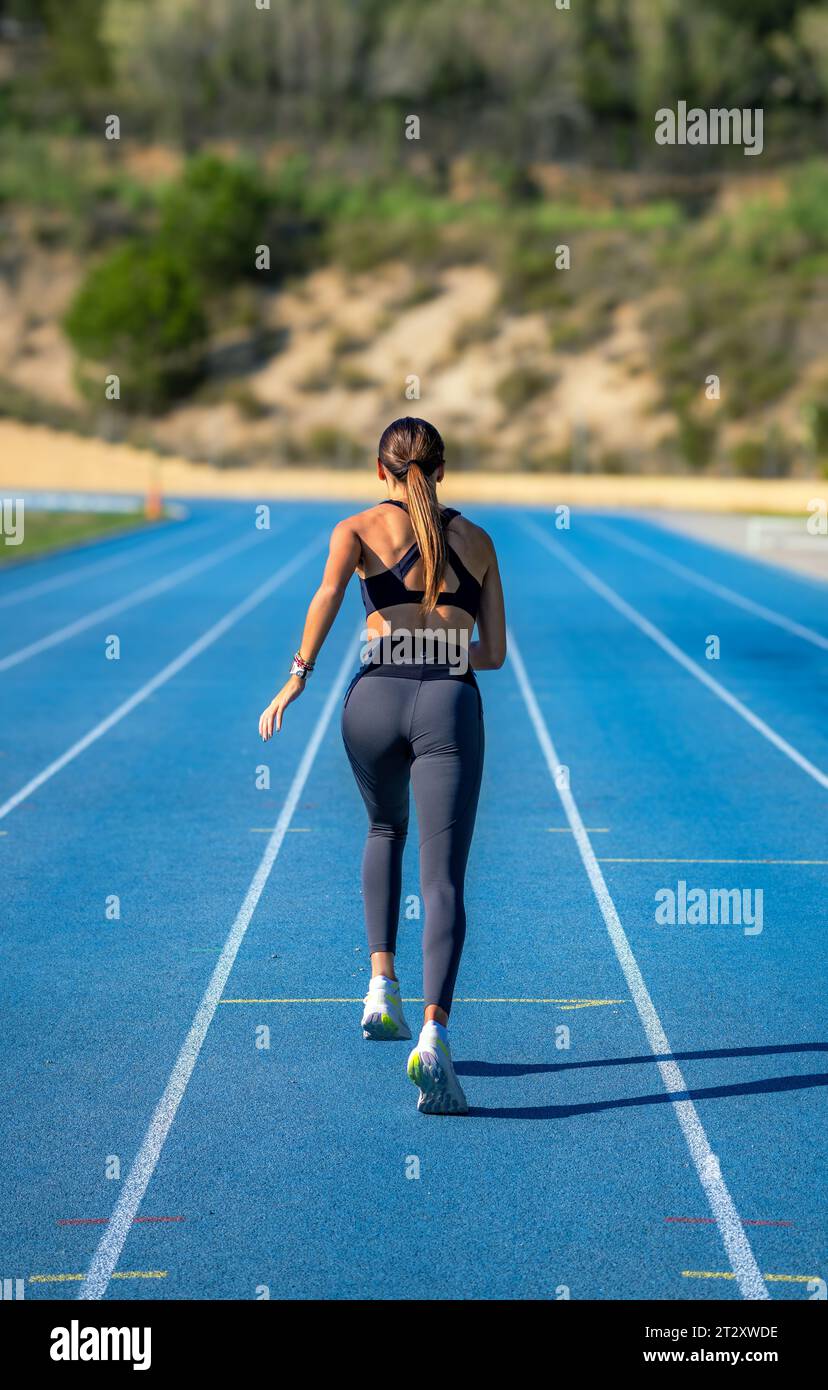 Schönes, gebräuntes, schlankes Läufermädchen, in enger Sportswear gekleidet, mit gebeugten Beinen und bereit, auf einer blauen Leichtathletikstrecke zu sprinten. Stockfoto