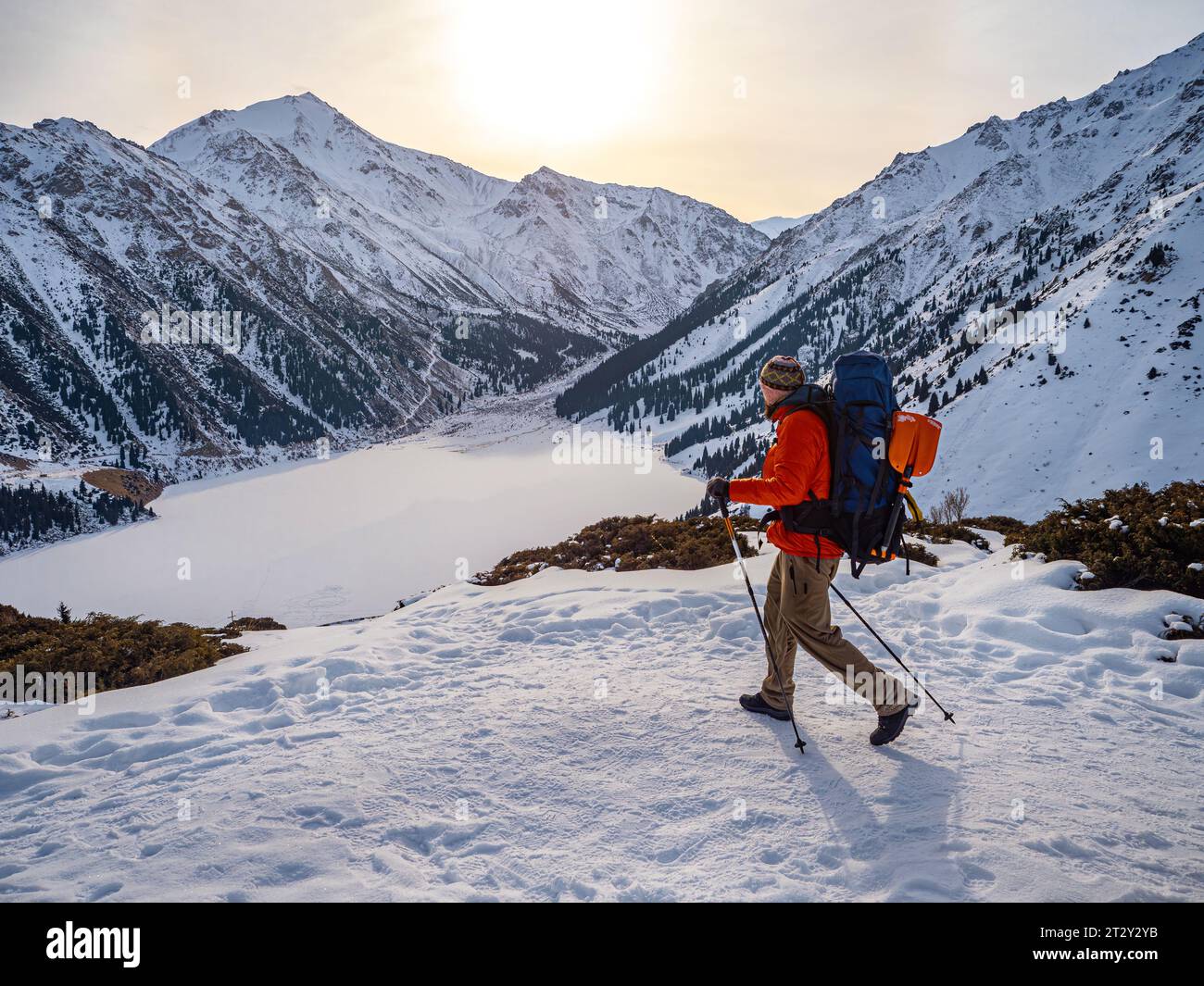 Ein Reisender macht im Winter Trekking hoch in den Bergen. Ein Mann in einer roten Daunenjacke mit Rucksack läuft vor dem Hintergrund einer gefrorenen mou Stockfoto