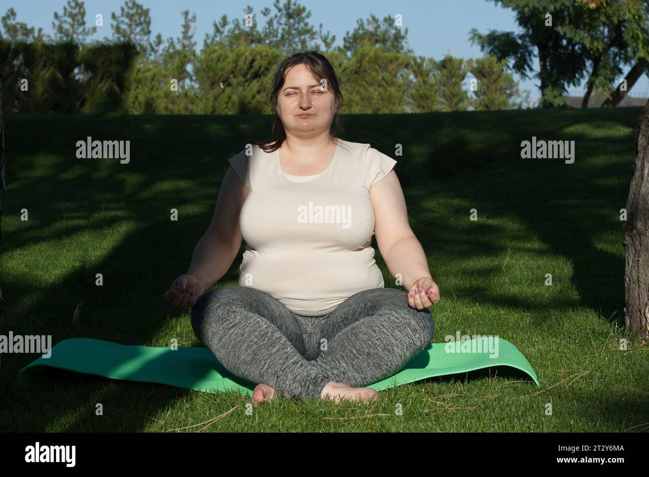 Porträt einer übergewichtigen kaukasischen Frau mittleren Alters, die Yoga in der Natur praktiziert. Fette Frau meditiert, entspannt in Lotusposition, sitzt auf Fitness Matte Stockfoto