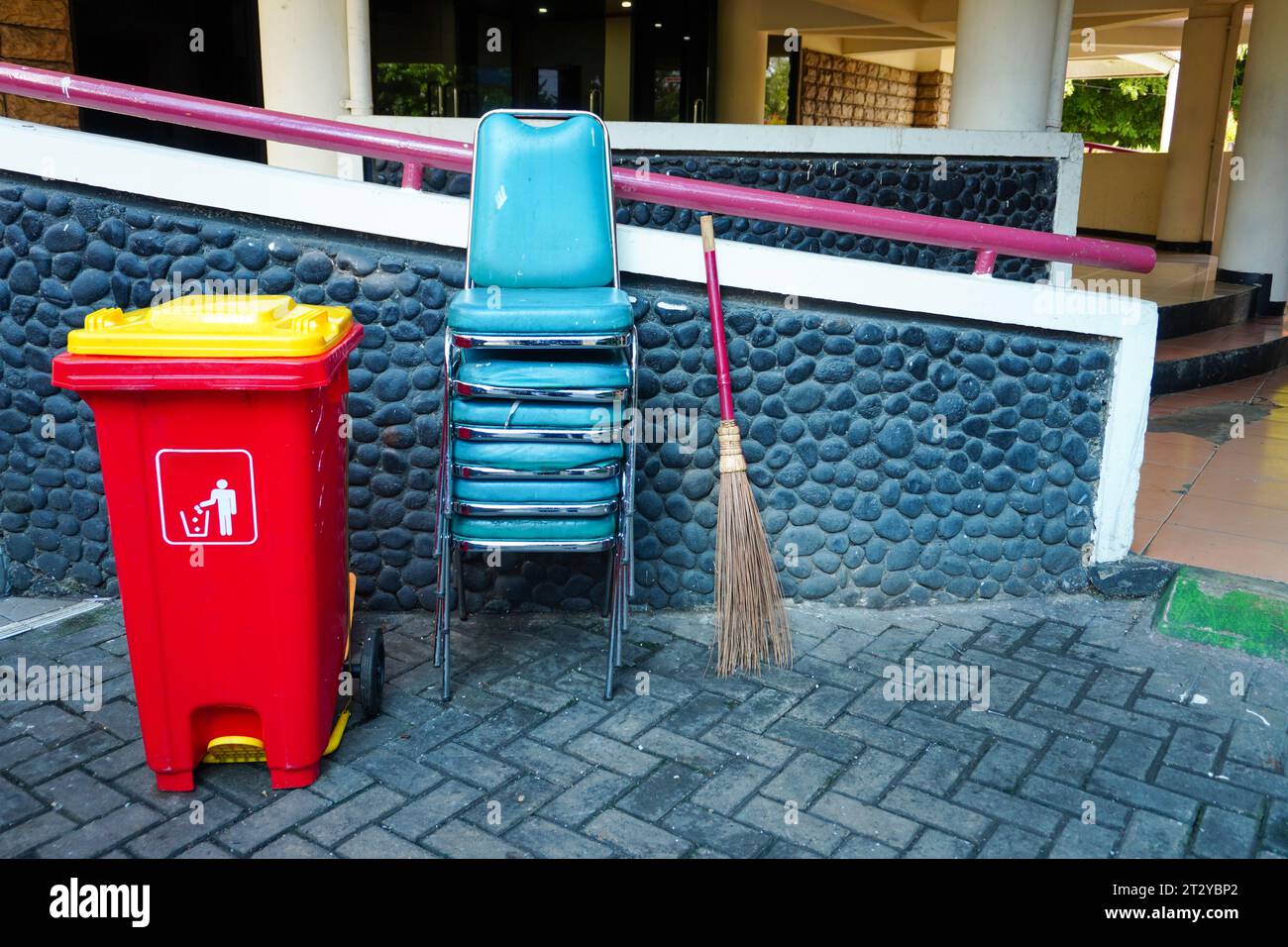 Grundausstattung des Hofes: Sapu ijuk, rote Plastikmüllbehälter im Freien und gestapelte Stühle Stockfoto