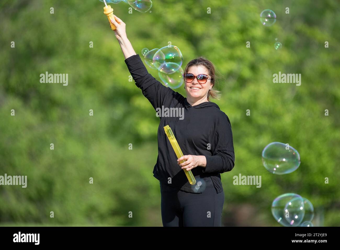 Schöne Frau mittleren Alters, die tagsüber mit Seifenblasen im Freien spielt. Sie schaut in die Kamera und lächelt. Das Konzept der Einfachheit im Leben Stockfoto