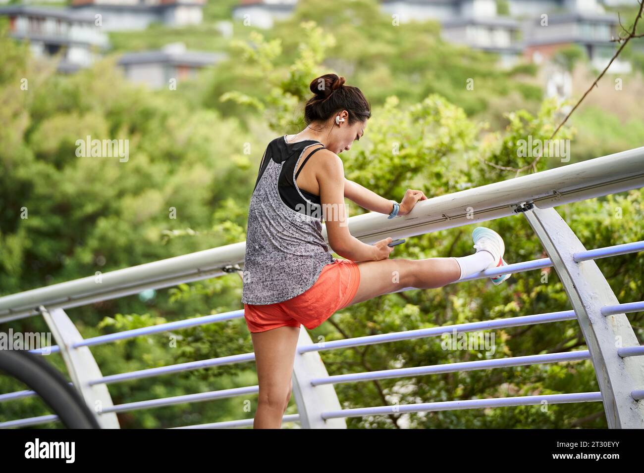 Junge asiatische Frau, die beim Aufwärmen auf das Handy schaut, indem sie draußen die Beine drückt Stockfoto