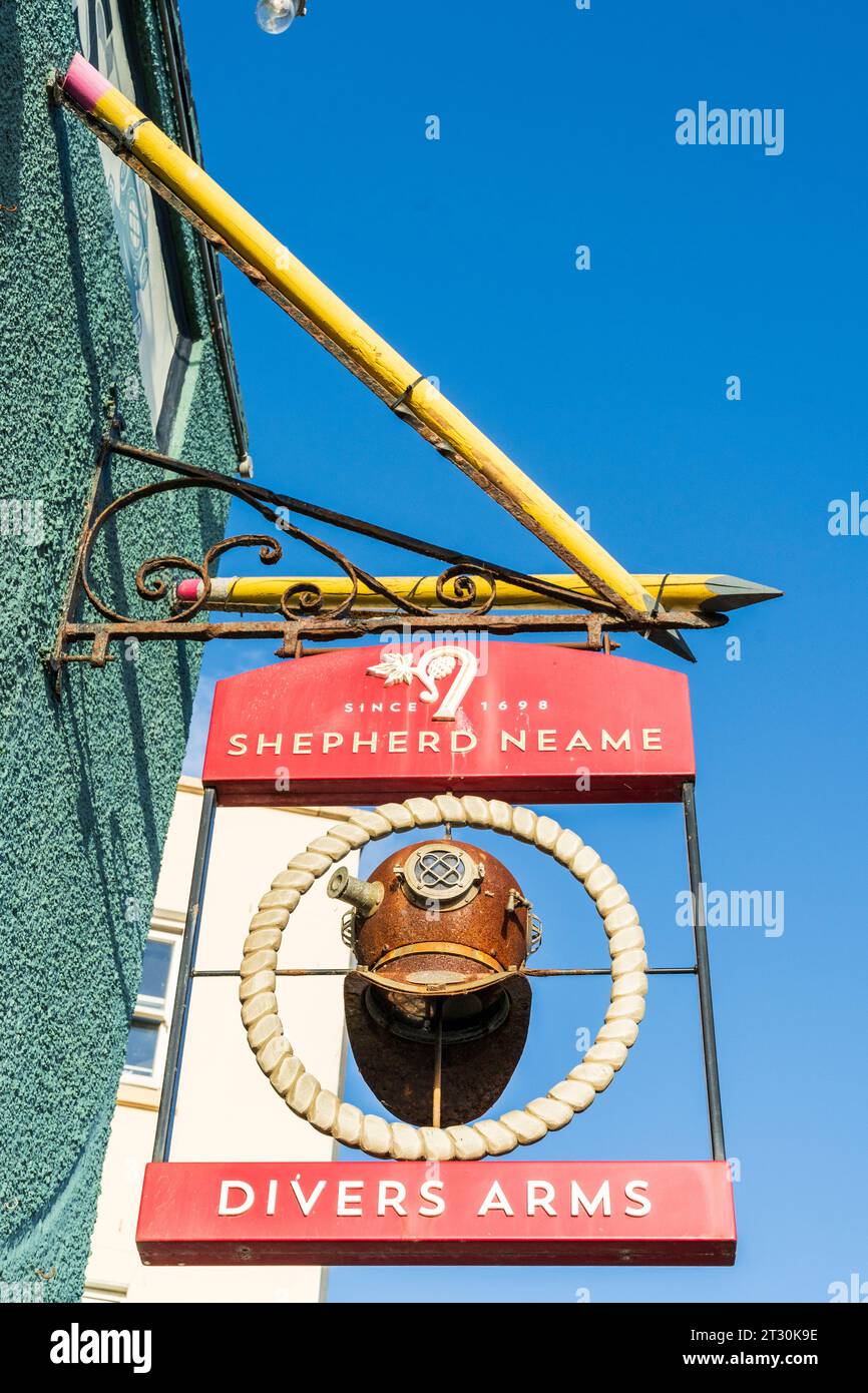 Ein typisches englisches Pub-Schild, das an einer Wand hängt, „The Divers Arms“, mit einem verrosteten Taucherhelm und dem Brauereinamen „Shepherd Neame“ Blue Sky. Stockfoto