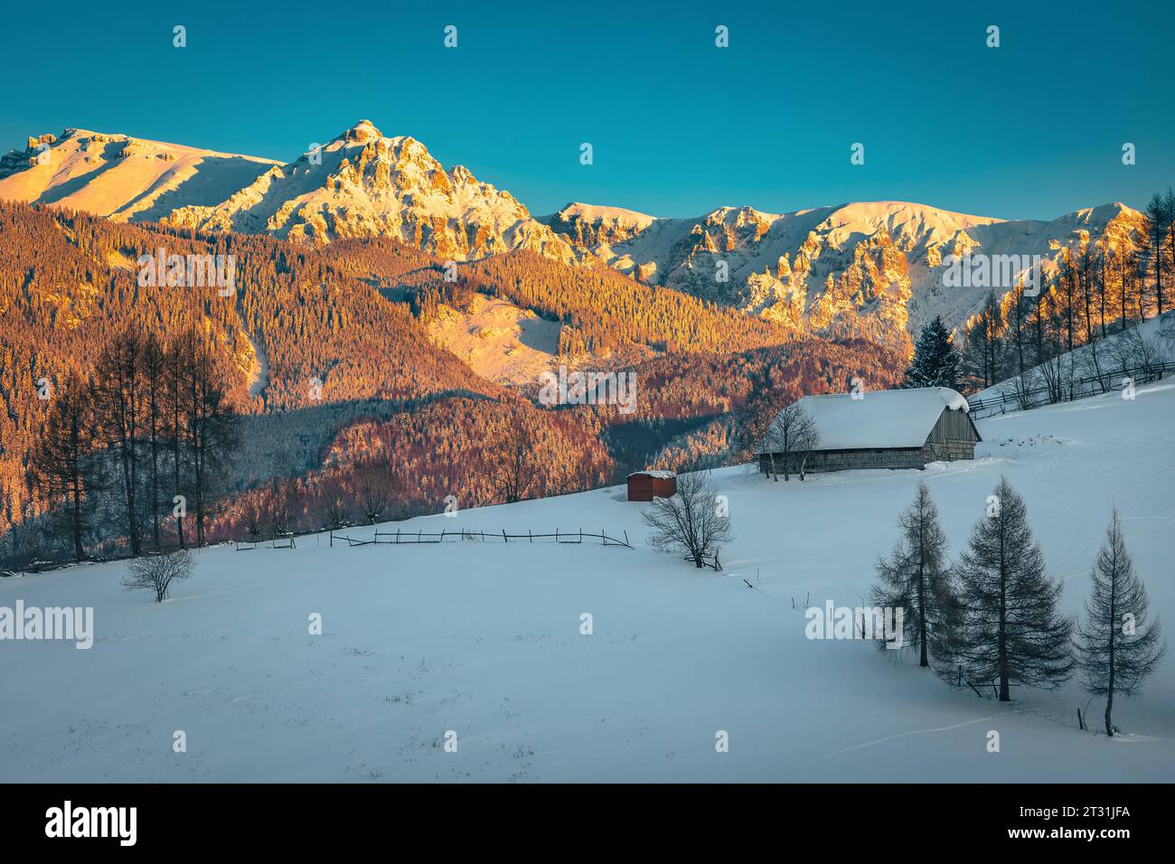 Wunderschöne Winterlandschaft mit schneebedeckter Holzhütte am Hang und majestätischen hohen Bergen im Hintergrund bei Sonnenuntergang, Bucegi Berge, Karpaten Stockfoto