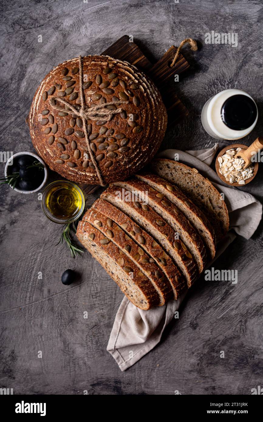 Gebackenes Sauerteigbrot aus Vollkornmehl und Kürbiskernen auf einem Gitter, Olivenöl und schwarze Olive auf einem rustikalen Holztisch. Handwerkliches Brot. Stockfoto