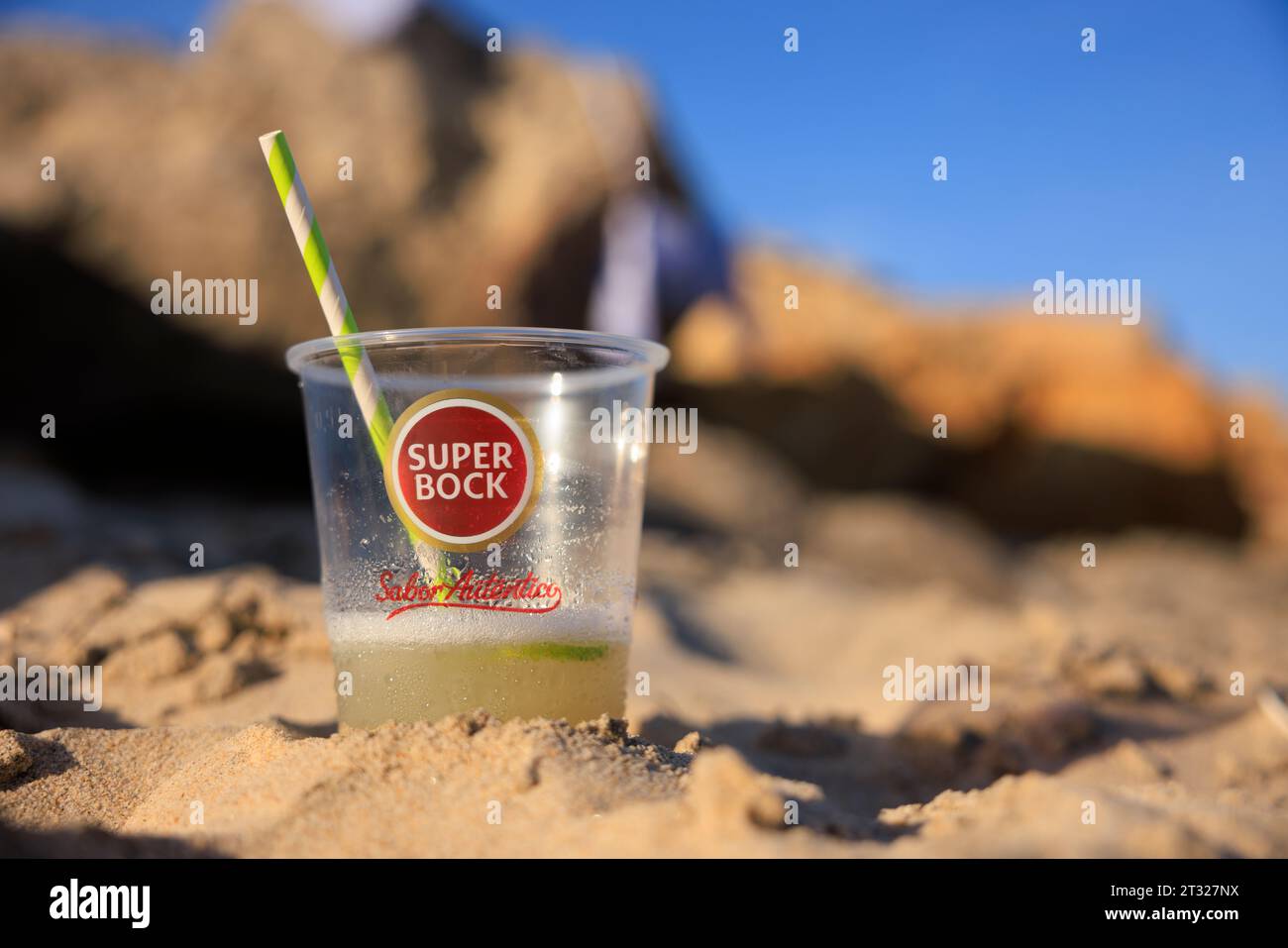 Ein Getränk in einem Super Bock Plastikglas im Sand an einem Strand auf Ilha do Farol (Leuchtturminsel) vor der Küste von Olhão in Portugal. Stockfoto