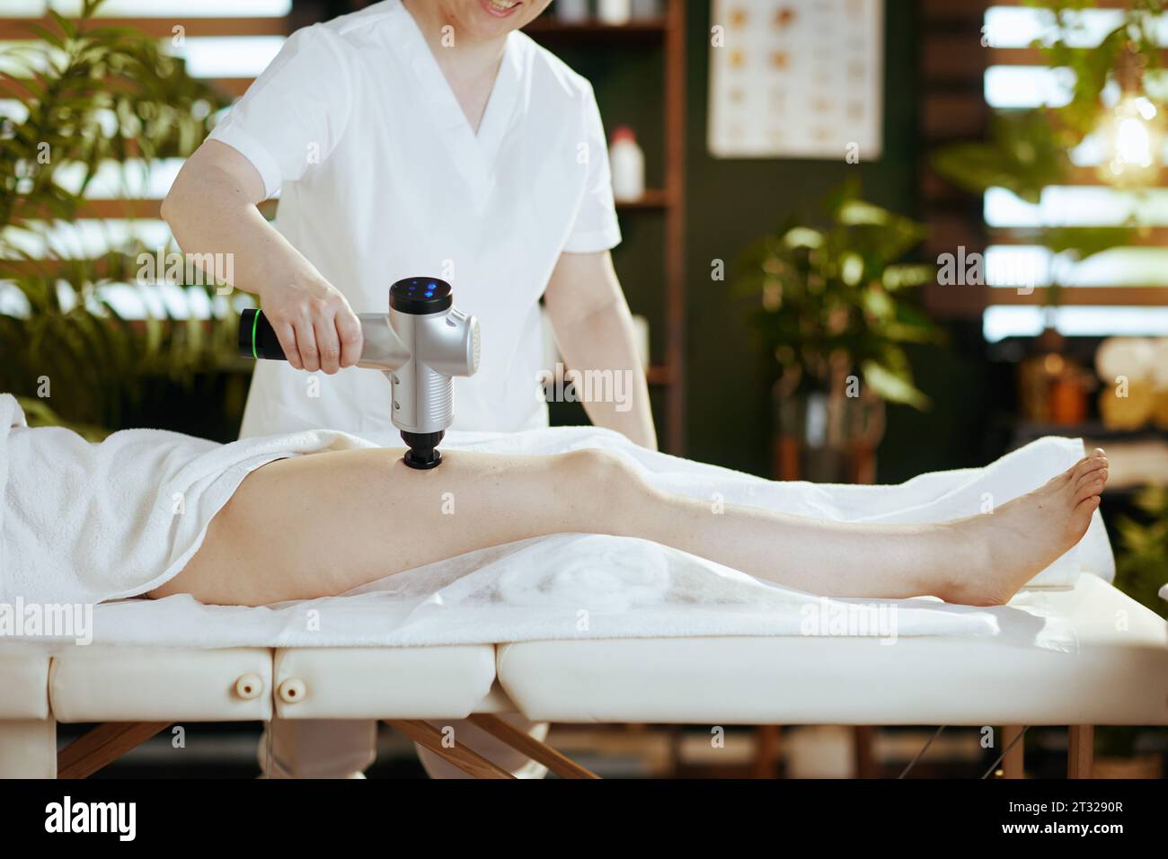 Gesundheitszeit. Nahaufnahme eines Massagetherapeuten in Massagekabine mit Massagepistole, der das Bein des Kunden auf dem Massagetisch massiert. Stockfoto