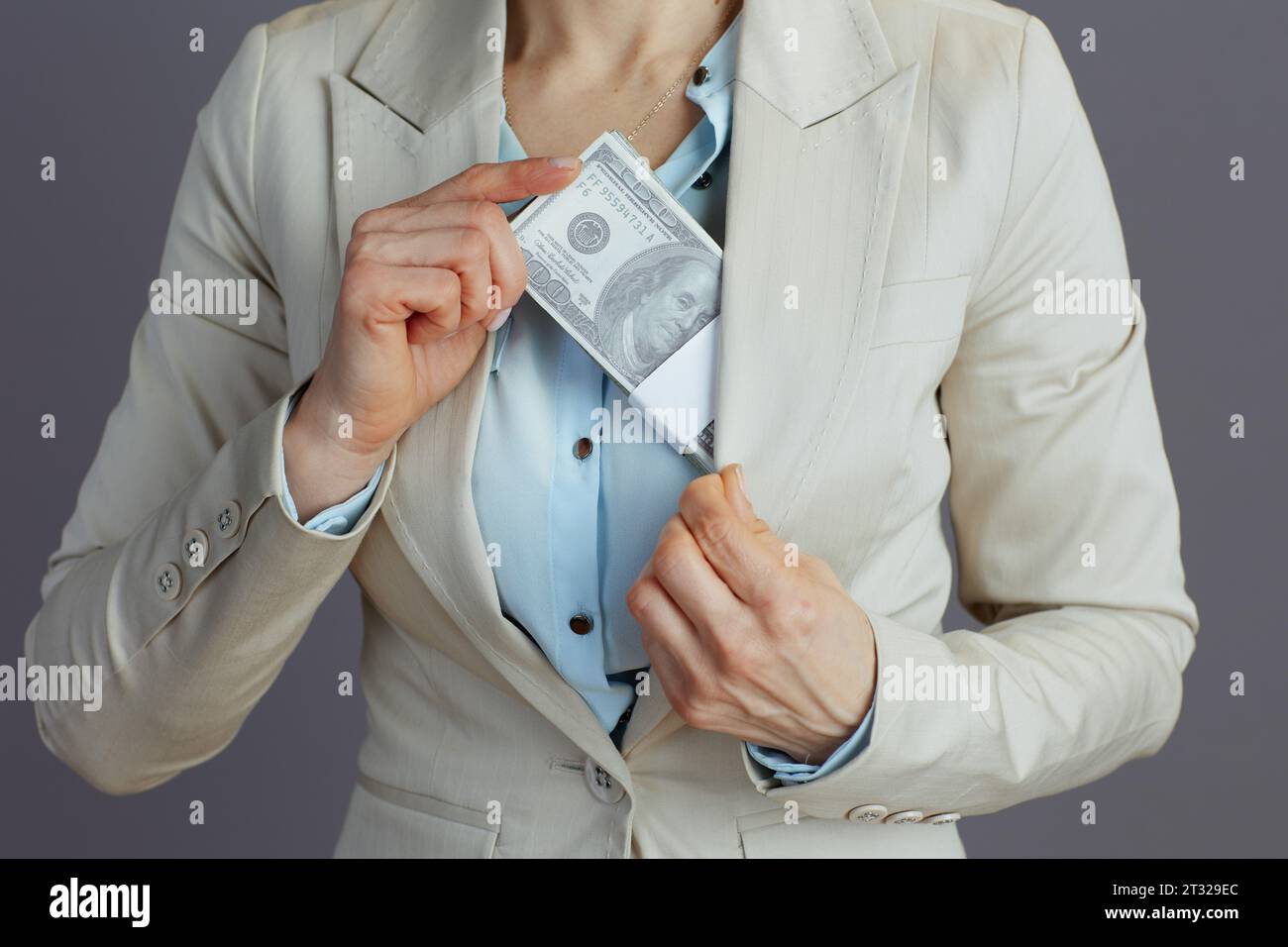 Nahaufnahme einer Geschäftsfrau in einem leichten Business-Anzug, die Dollargeldpackungen vor grauem Hintergrund versteckt. Stockfoto