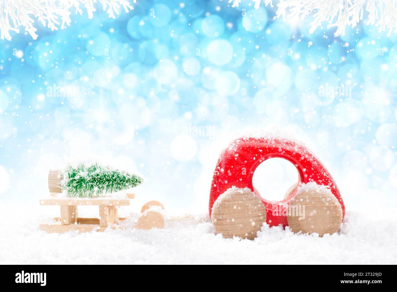 Rotes hölzernes Auto, das Schlitten mit grünem Weihnachtsbaum in der Perspektive auf hellblauem Bokeh-Winterhintergrund mit weißen schneebedeckten Zweigen und fallendem Schnee zieht Stockfoto