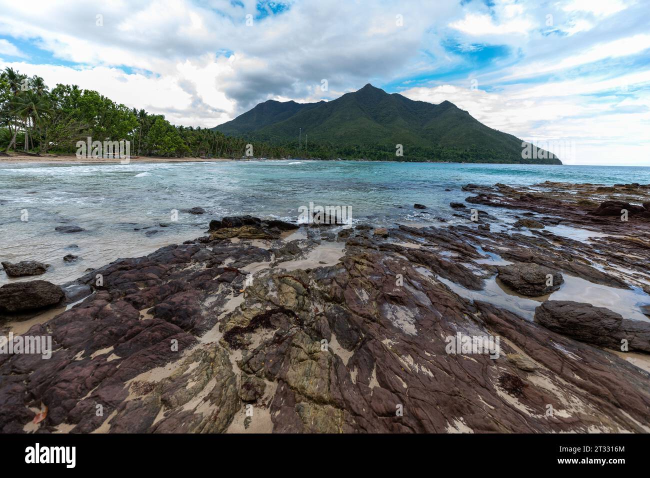 Die felsige Küste des wunderschönen tropischen Strandes blickt auf Berge, die von üppigem grünen Dschungel bedeckt sind Stockfoto