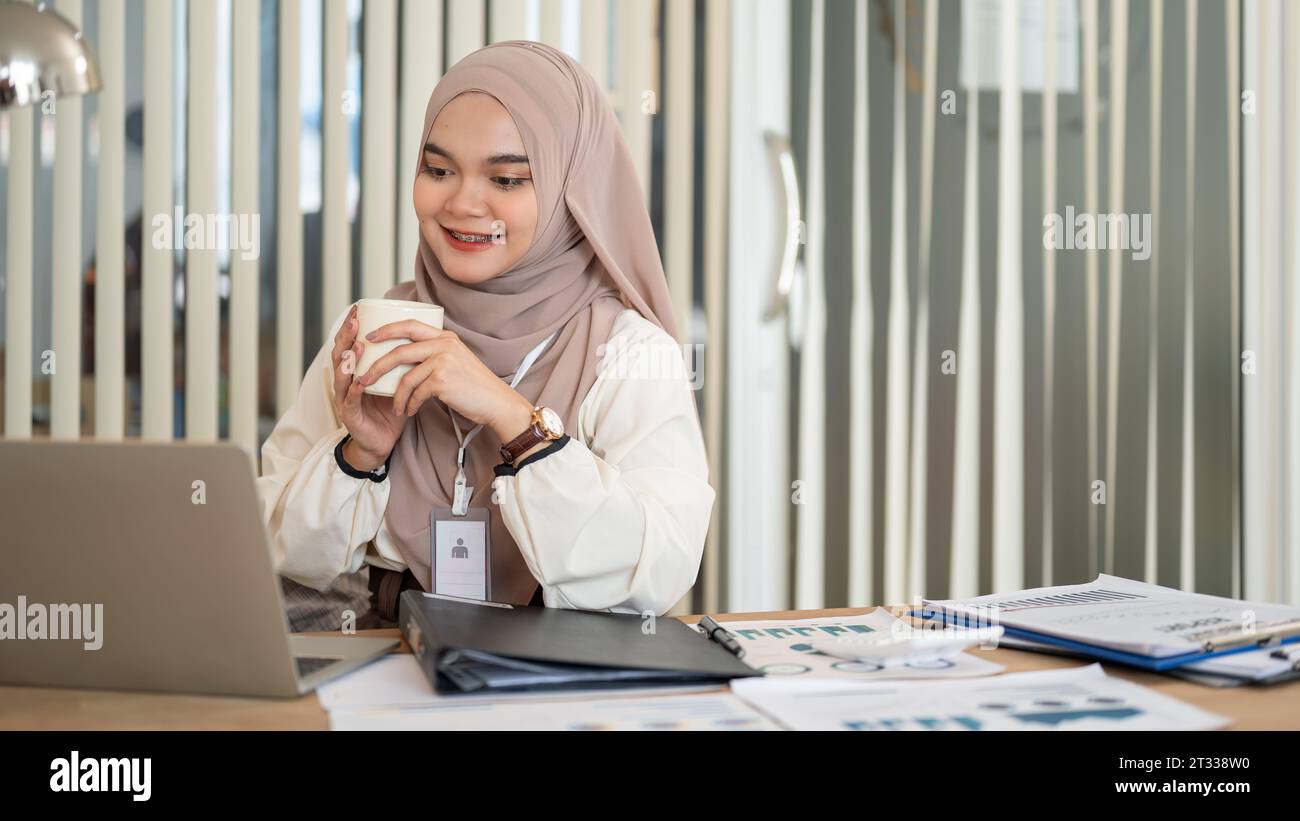 Eine fröhliche und attraktive Millennials asiatisch-muslimische Geschäftsfrau trinkt ihren Morgenkaffee, während sie an ihrem Schreibtisch im Büro an ihrem Laptop arbeitet. Stockfoto Eine fröhliche und attraktive Millennials asiatisch-muslimische Geschäftsfrau trinkt ihren Morgenkaffee, während sie an ihrem Schreibtisch im Büro an ihrem Laptop arbeitet. Stockfoto